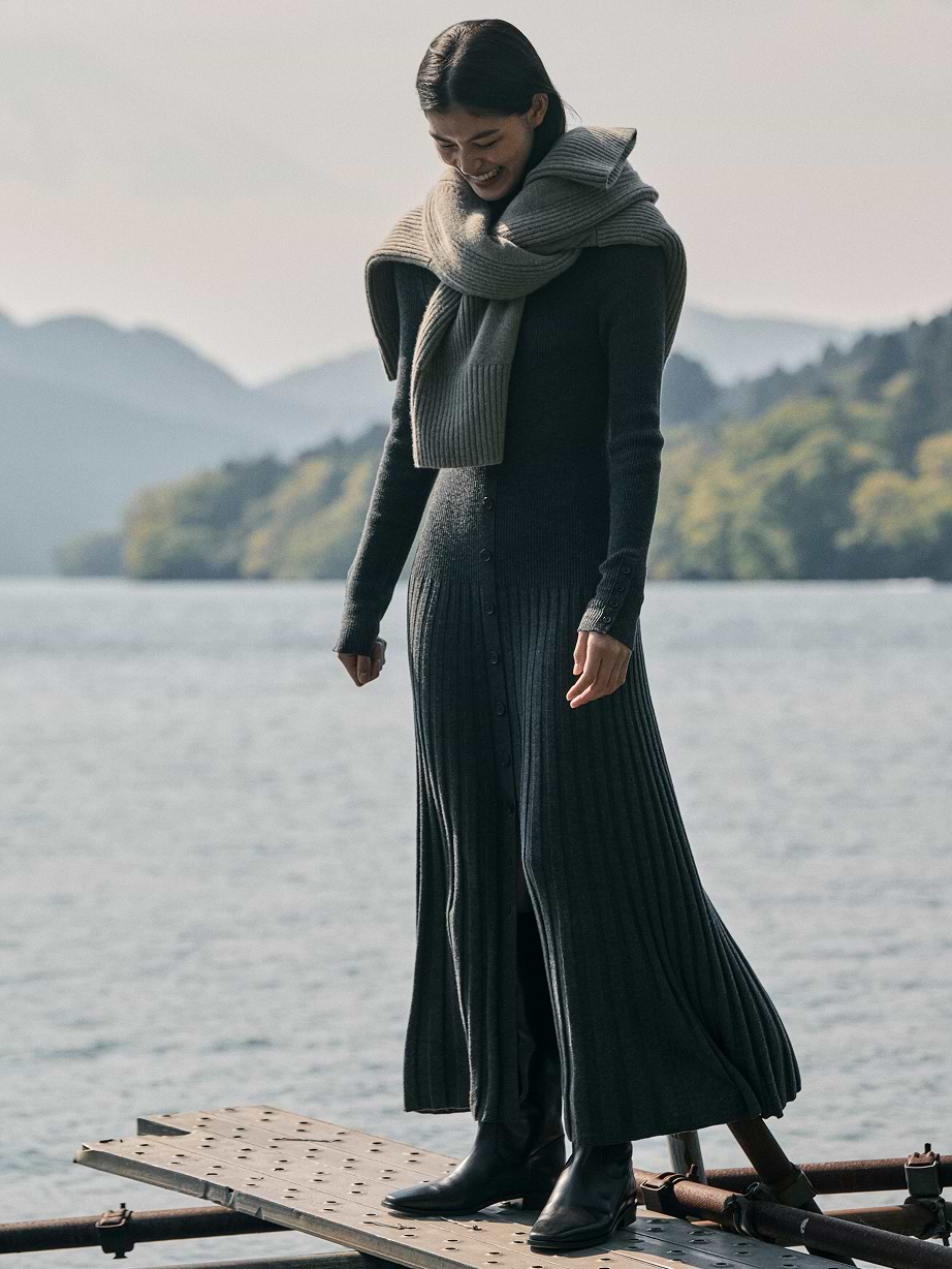 Smiling person in a long gray ribbed sweater dress and scarf stands on a lakeside dock with mountains in the background.