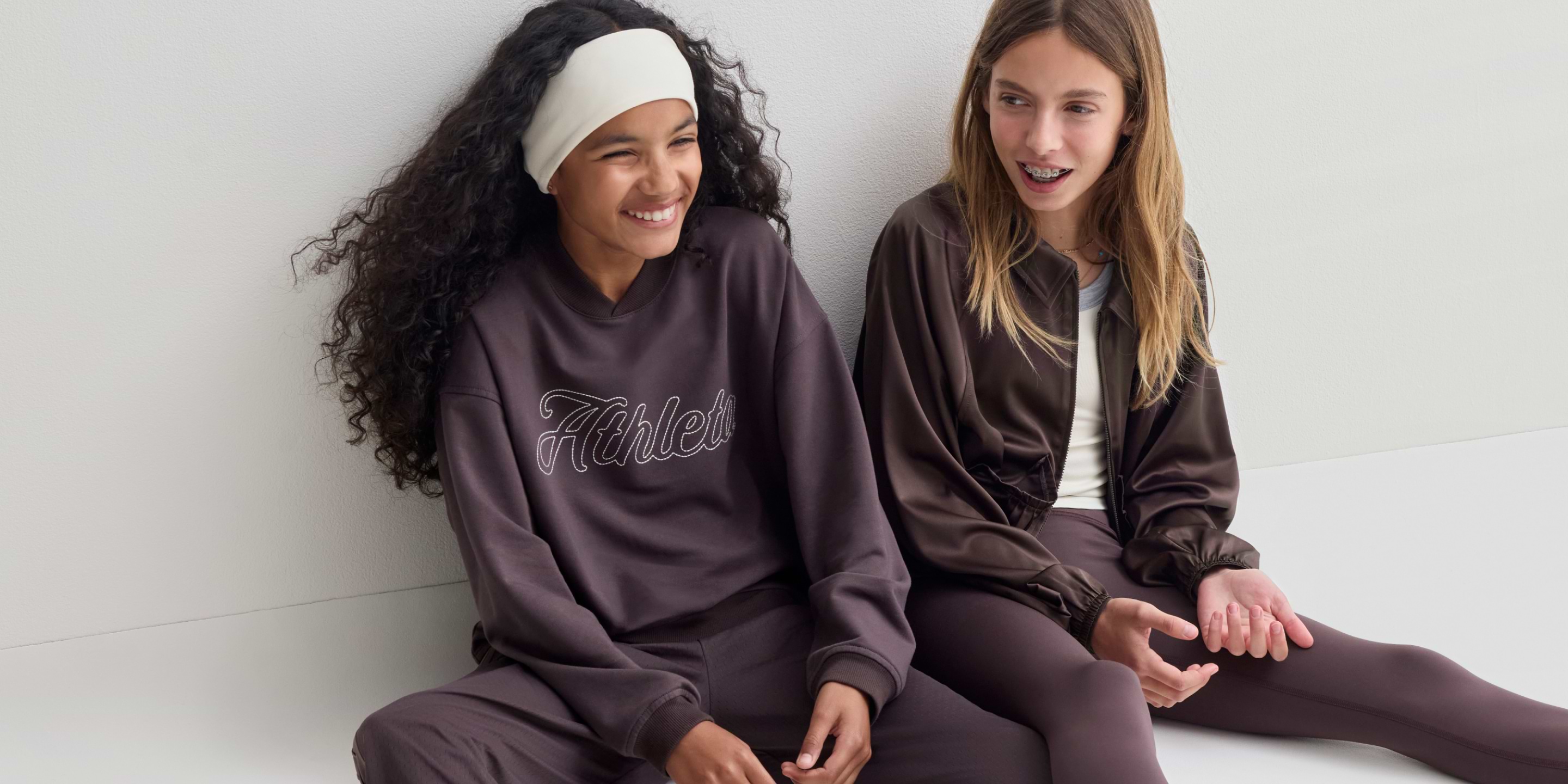 Two young girls in brown athleisure sit against a white wall, smiling; one wears a headband and a sweatshirt reading “Athleta,” the other a jacket and leggings.