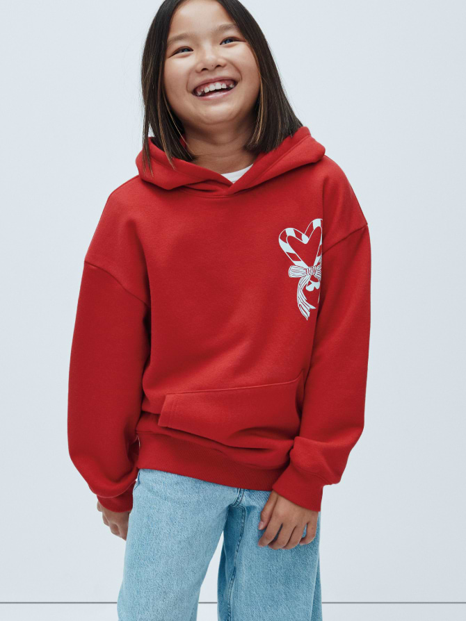 Smiling child in a red hoodie with a white candy-cane heart graphic, wearing light blue jeans against a plain background.