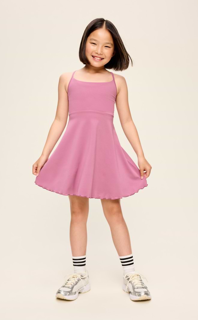 Smiling child in a pink sleeveless dress holding out the skirt, wearing striped socks and sneakers on a plain studio background.