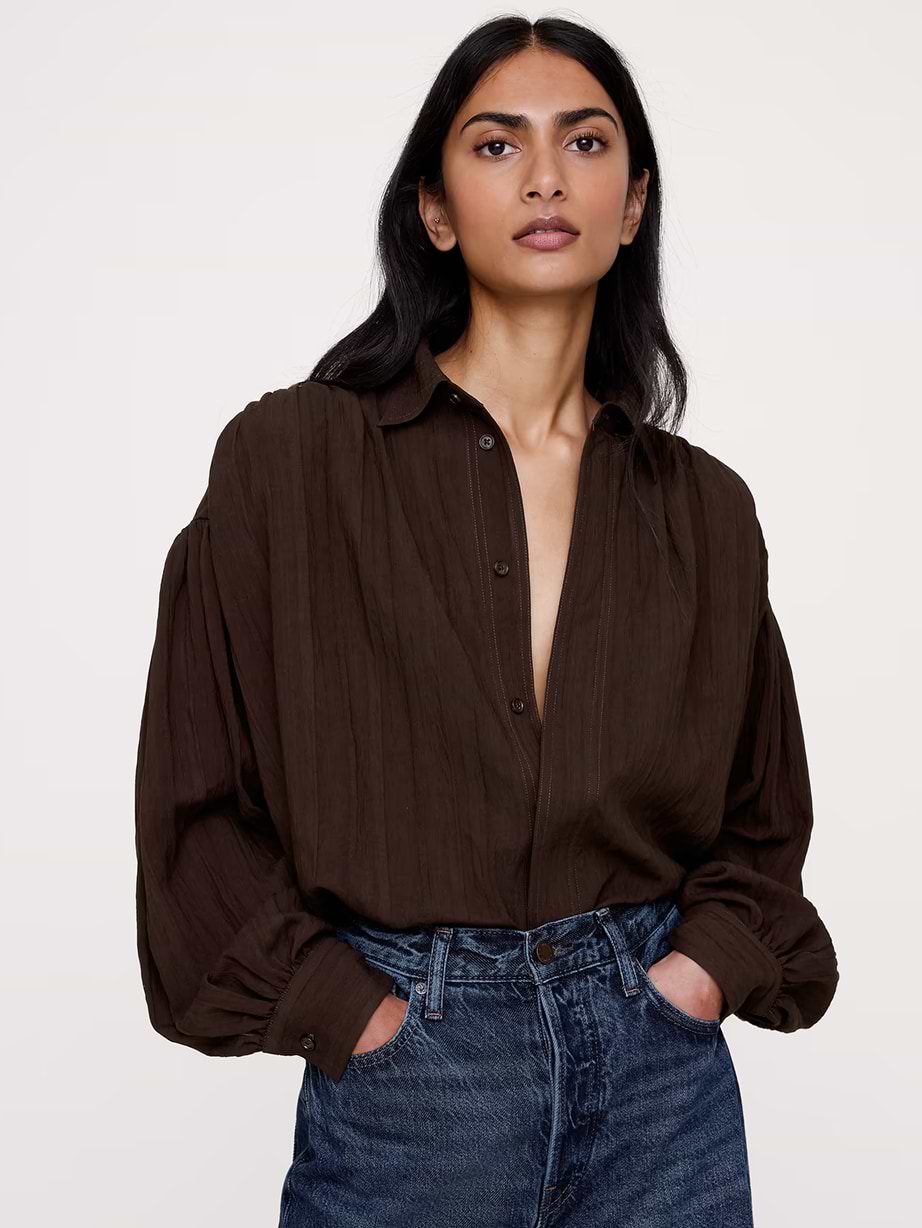 Person wearing a loose dark brown button-up shirt and high-waisted blue jeans, hands in pockets, against a plain background.