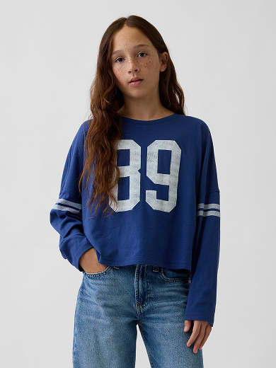 Person wearing a blue long-sleeve top with “89” and jeans against a plain background.