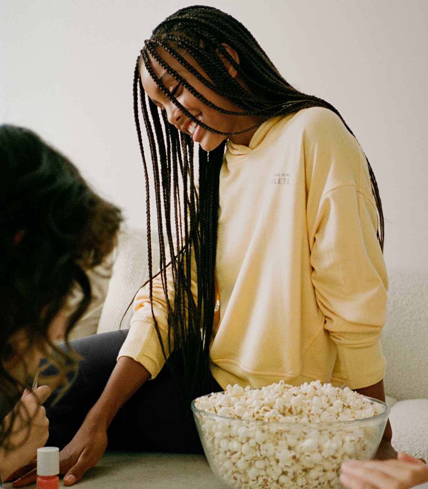 Smiling girl with long braids in a yellow hoodie leans over a large bowl of popcorn.