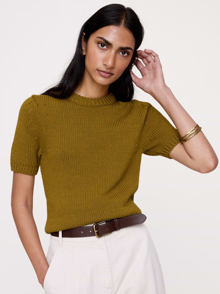 Person wearing an olive short‑sleeve knit sweater with white trousers and a brown belt.