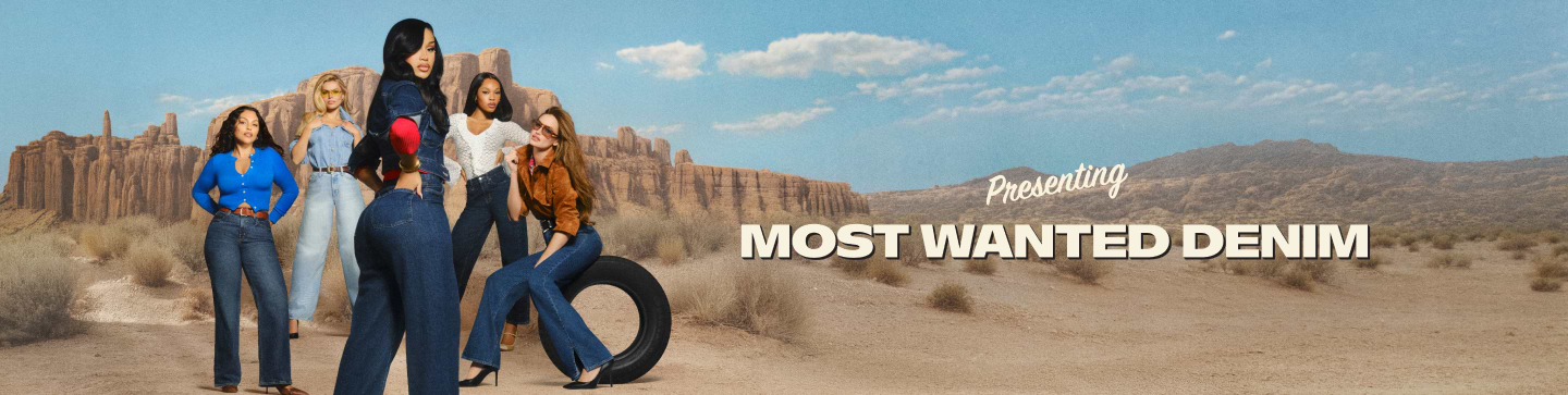 Six women in denim outfits pose in a desert with rocky cliffs and scrub under a blue sky; one sits on a tire.