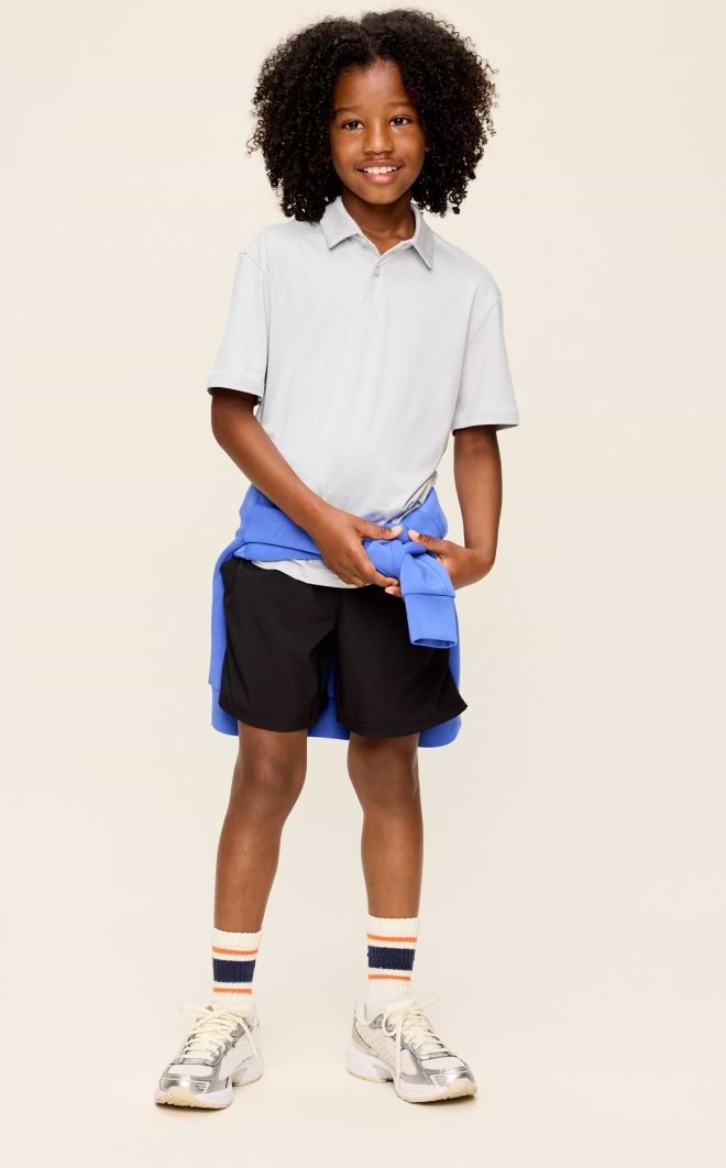 Smiling child in a light gray polo and black shorts, blue sweatshirt tied at waist, striped socks and sneakers.