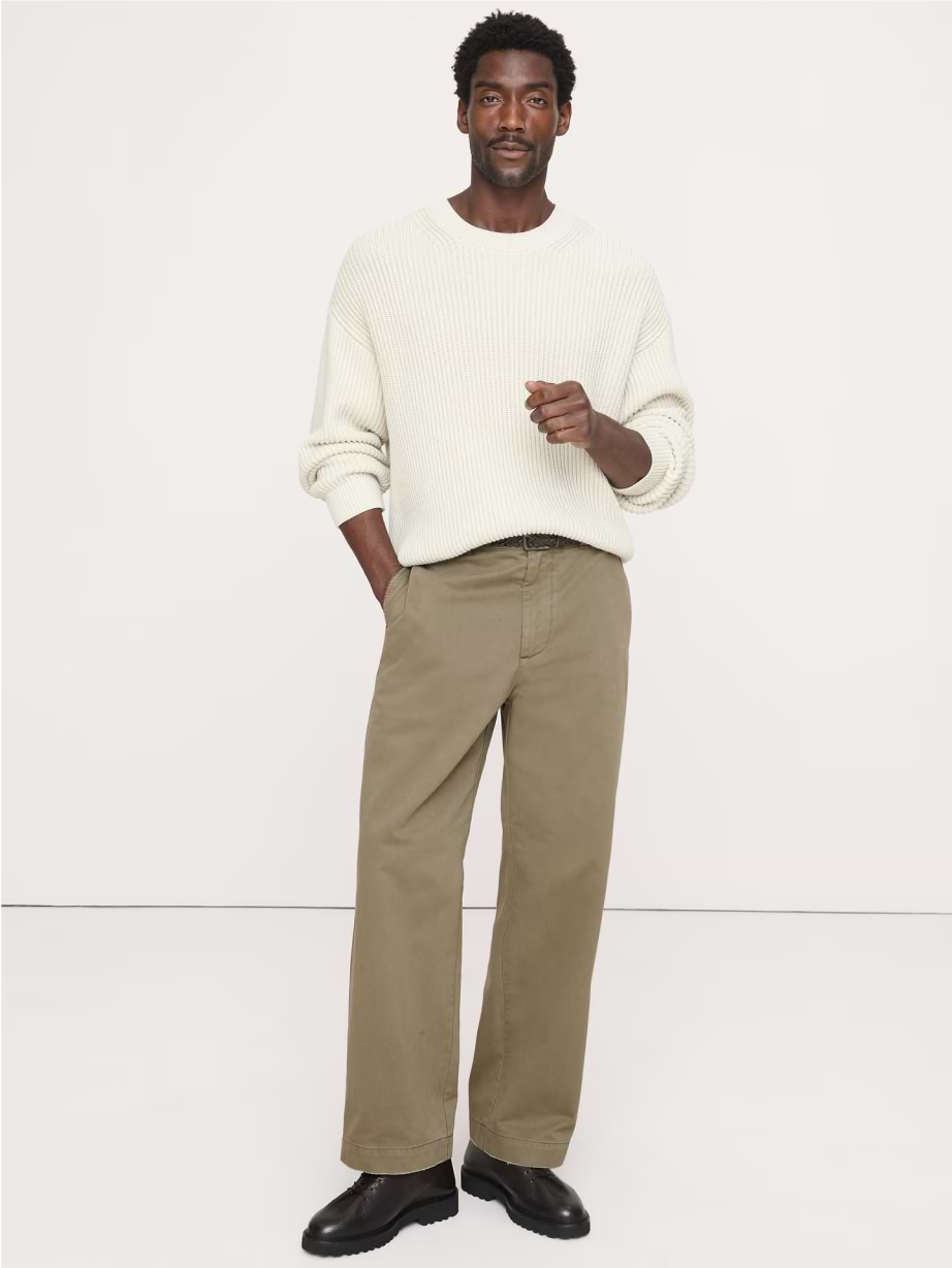 Person in a cream knit sweater, olive wide-leg pants, and black shoes standing against a plain background with one hand in a pocket.