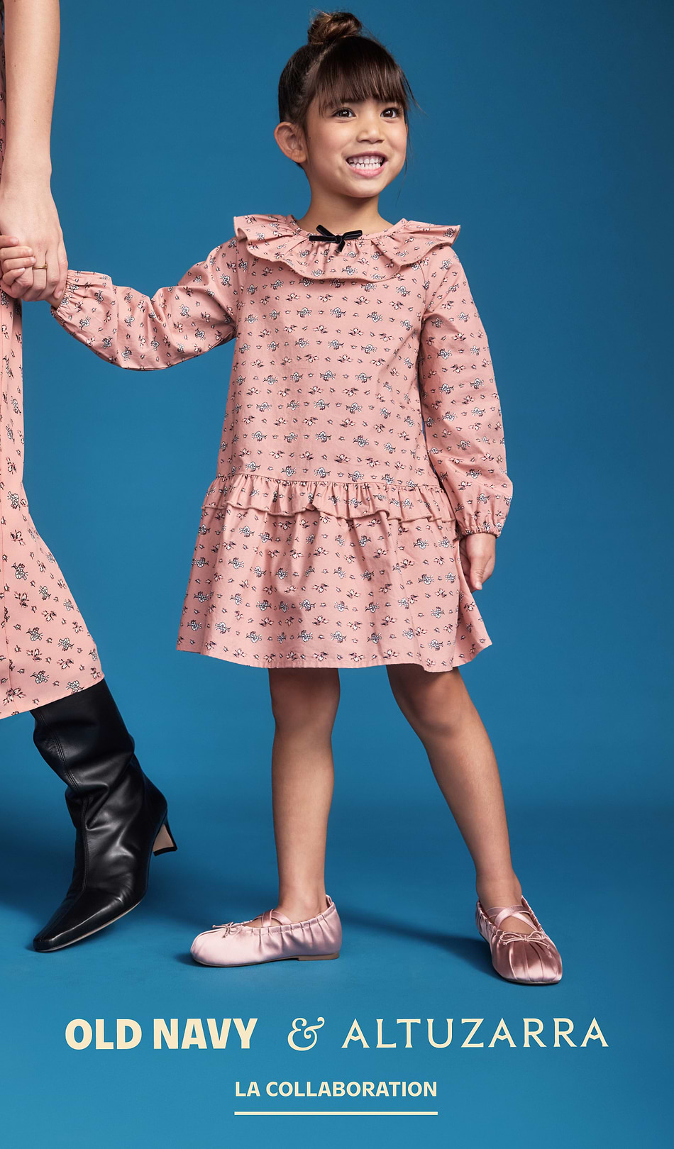 Smiling child in a pink floral ruffle dress and ballet flats holds an adult’s hand; text reads “Old Navy & Altuzarra – Shop the Collab.”