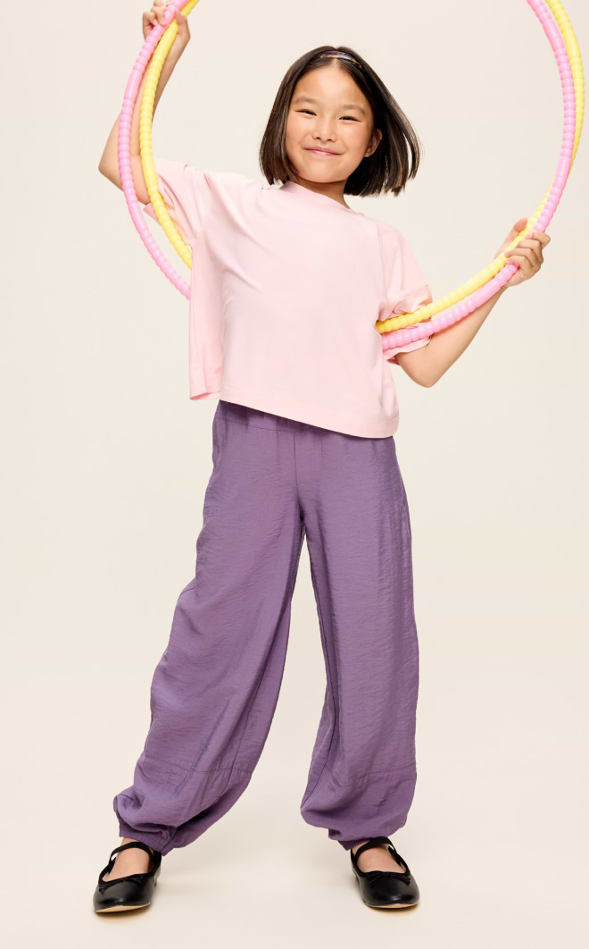 Smiling child holding a pink-and-yellow hula hoop, wearing a light pink T-shirt, loose purple pants, and black shoes on a plain background.