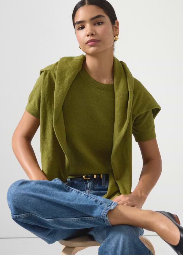 Person sitting cross‑legged on a stool wearing an olive‑green knit top with a matching sweater over the shoulders, blue jeans, and black flats against a plain light background.