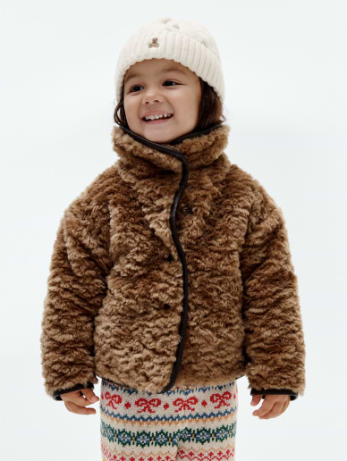 Smiling child in a fuzzy brown jacket, cream knit beanie, and colorful patterned pants against a plain white background.