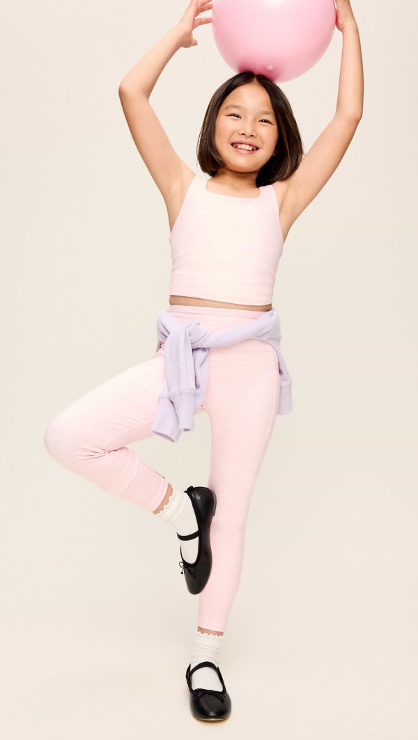 Smiling child balancing on one leg in pink activewear, holding a pink ball overhead.