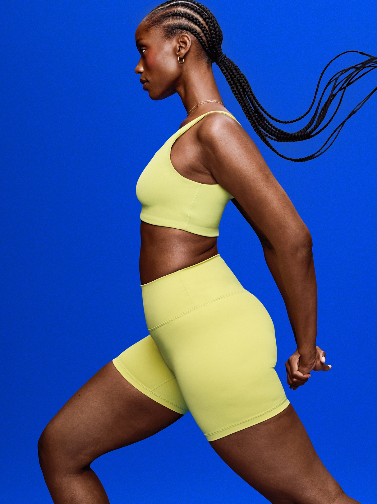 Side-profile athlete with long braids in lime-green sportswear, mid-stride against a bright blue background.