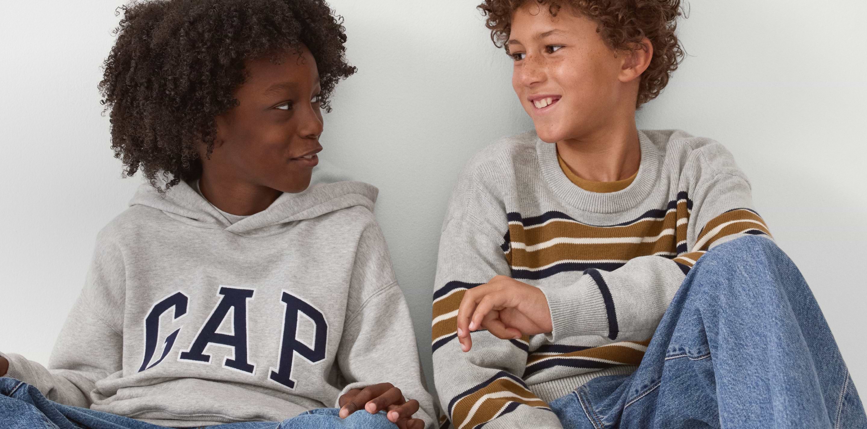 Two kids sit smiling at each other; one wears a gray GAP hoodie, the other a gray striped sweater and jeans.