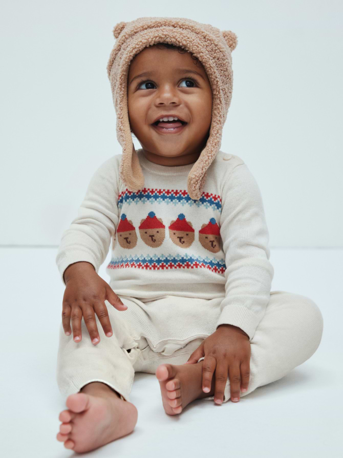 Smiling toddler in a fuzzy bear-ear hat and cream sweater with acorn pattern, sitting barefoot.