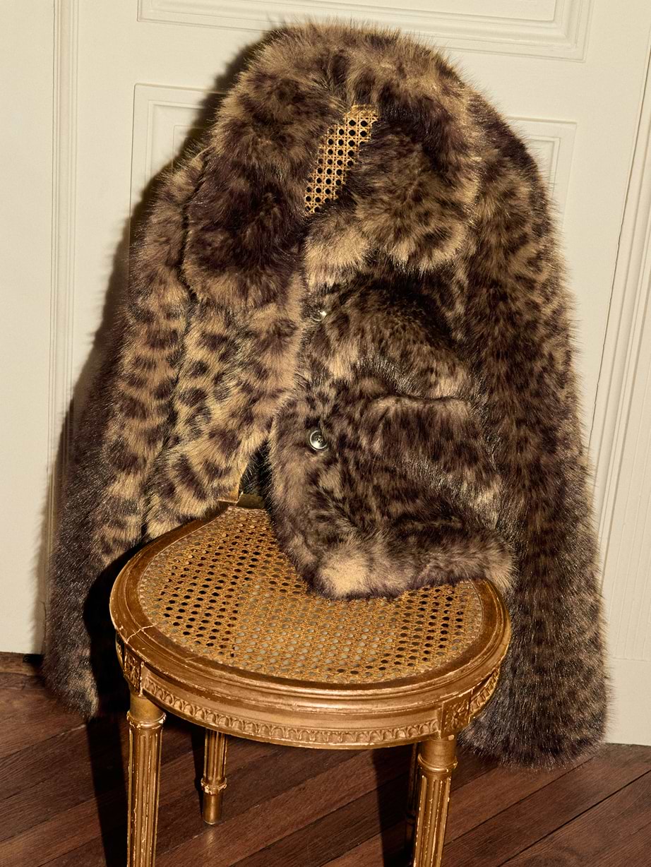Leopard-print fur coat draped over a gilded cane-seated chair on a wooden floor, against a white paneled wall.