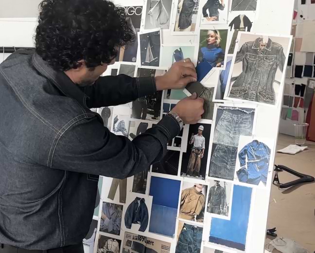 Person in a denim shirt arranging fabric swatches on a mood board covered with denim clothing photos in a studio.