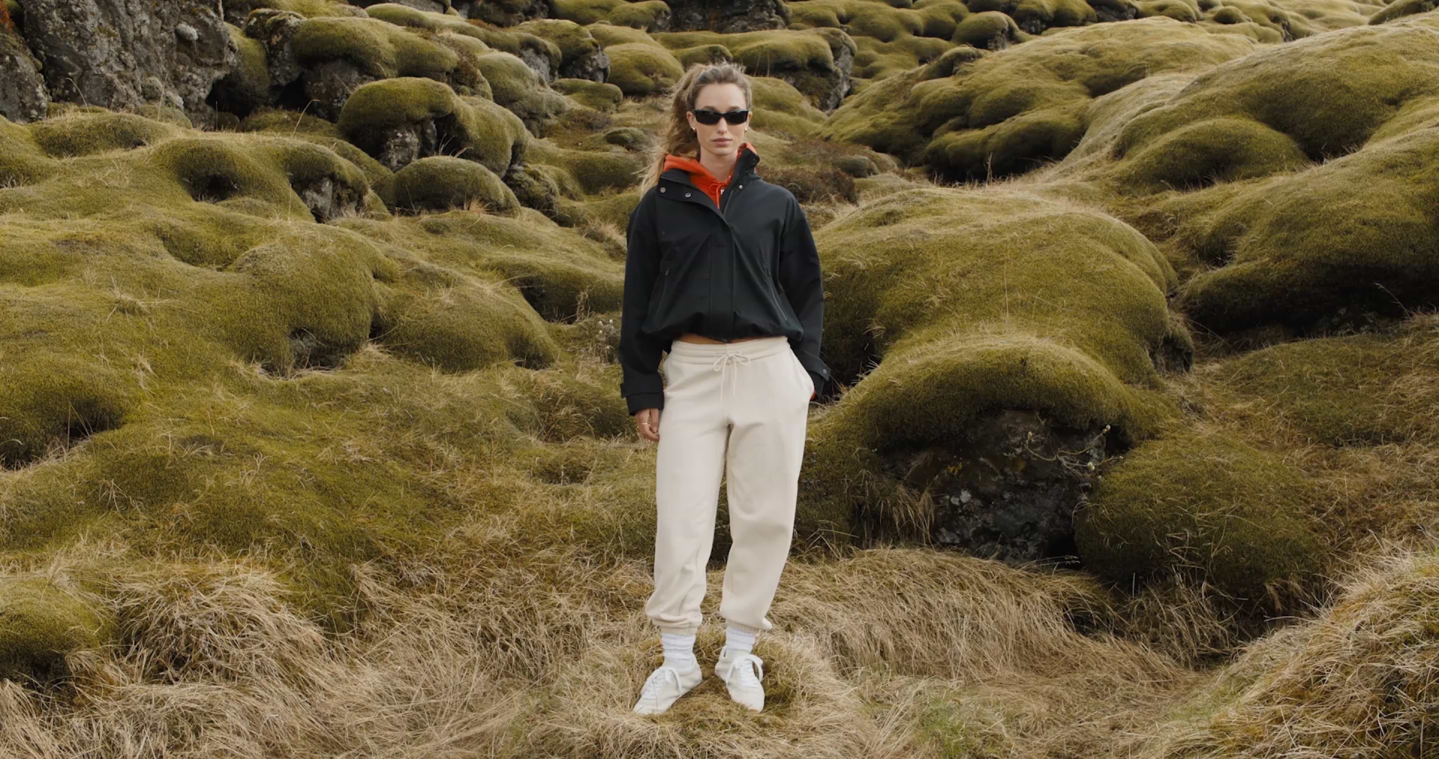 Person in a black jacket and beige joggers stands on a moss-covered rocky field, wearing sunglasses and white sneakers.