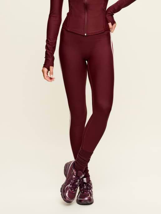 Cropped view of person wearing burgundy leggings and zip jacket with matching sneakers, standing against a light background.