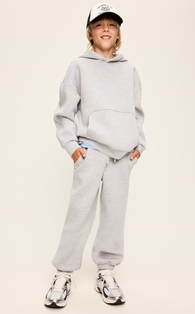 Smiling child in gray hoodie and sweatpants, wearing a cap and sneakers, against a plain background.