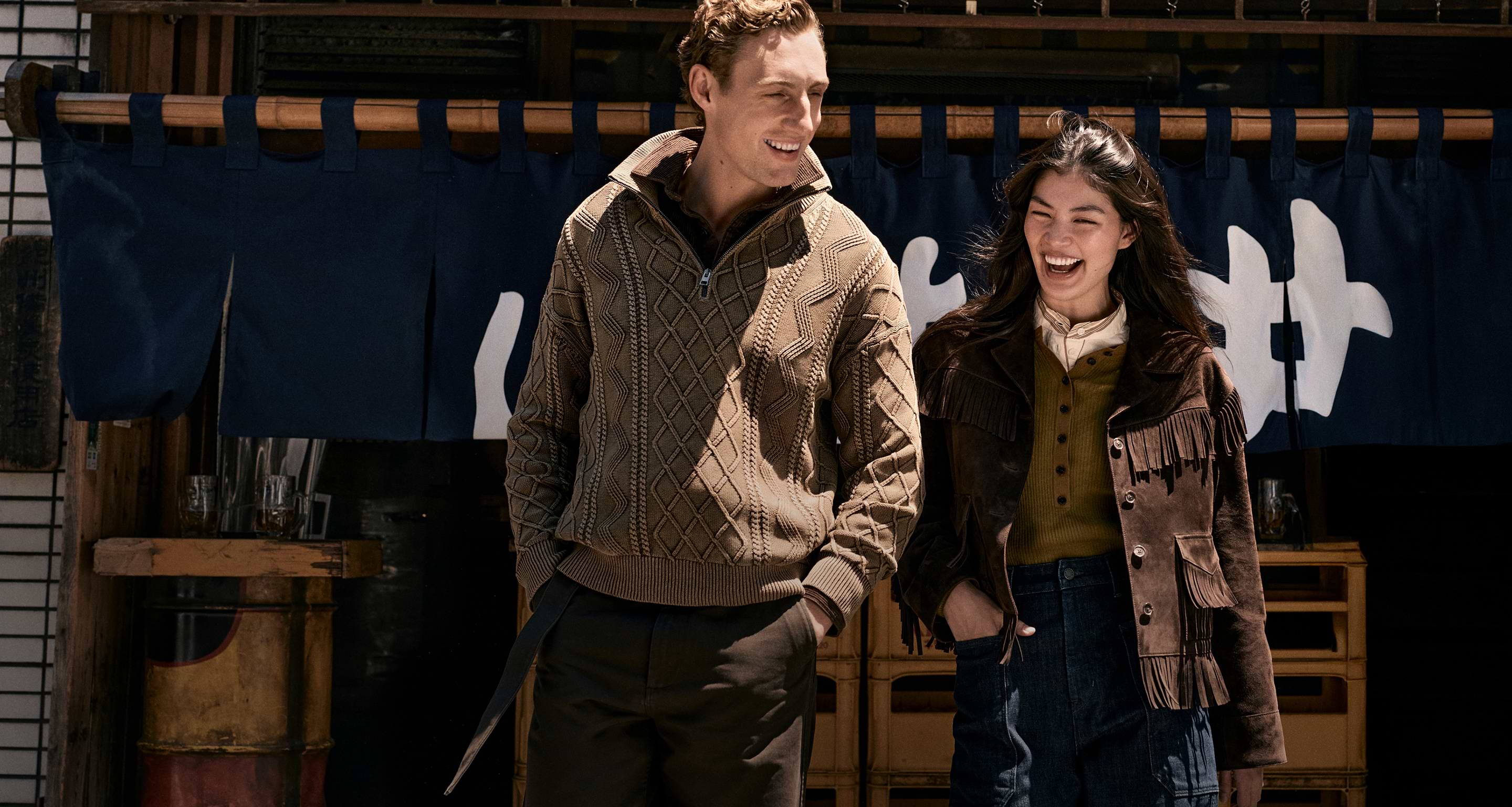 Two people smile while walking side by side—one in a tan cable-knit sweater, the other in a brown fringed jacket—against a blue curtain with white characters.