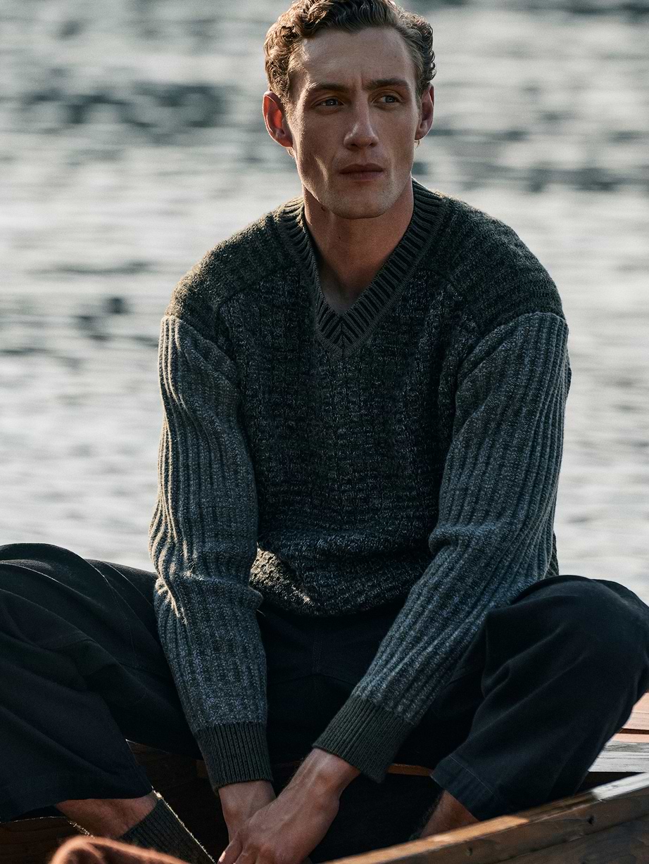 Person in a gray ribbed V-neck sweater sits in a wooden boat by the water, looking off to the side.