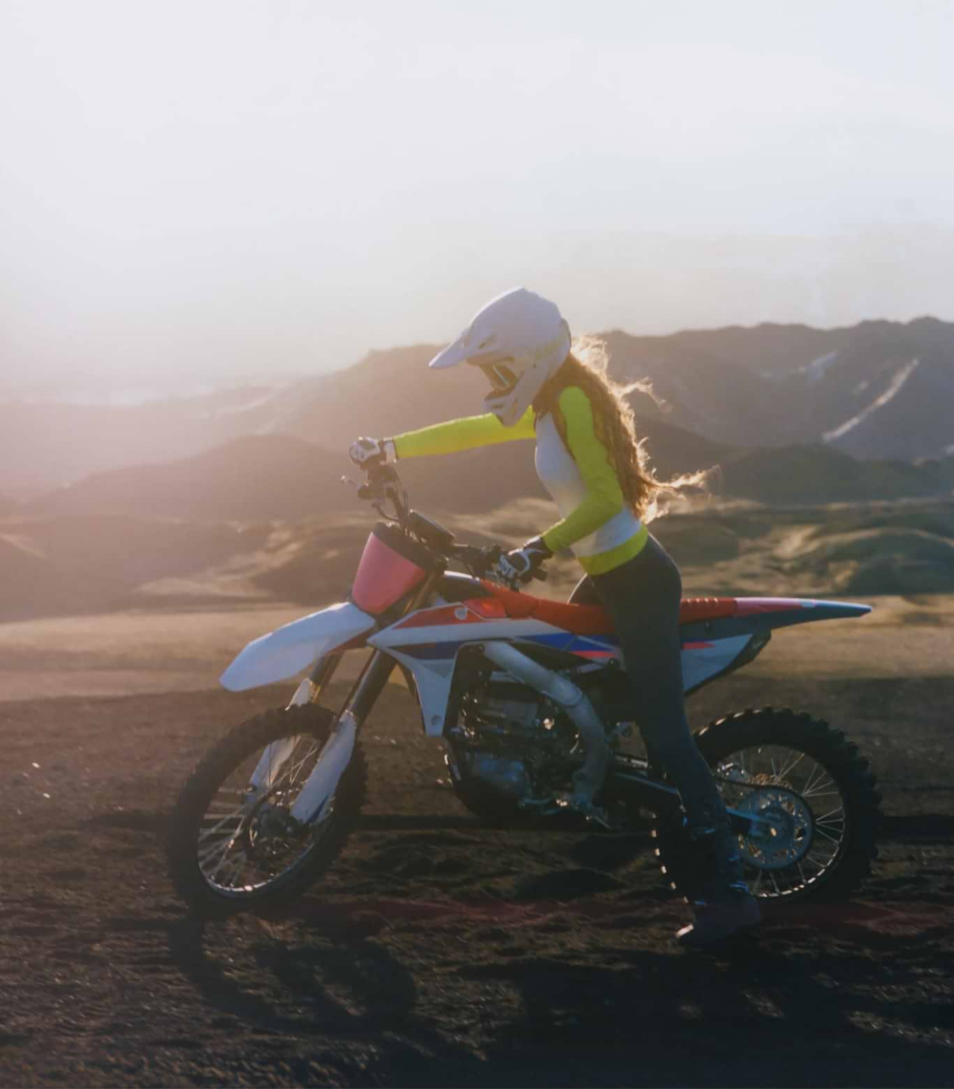 Helmeted rider on a dirt bike at sunset in a mountainous landscape.