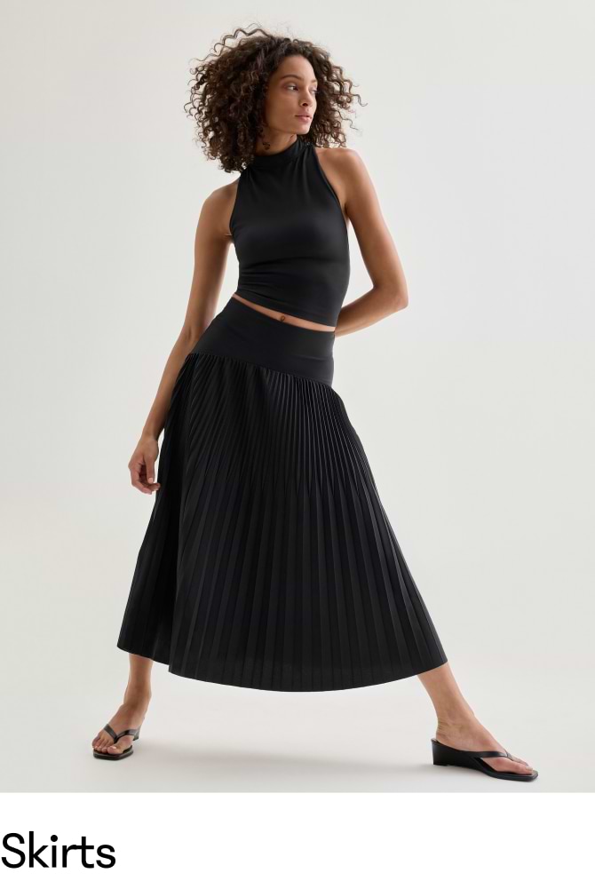 Woman wearing a black sleeveless crop top, black pleated midi skirt, and sandals, posing in a studio.