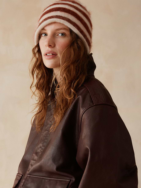 Person wearing a fuzzy red-and-cream striped beanie and a dark brown leather jacket, looking at the camera against a beige backdrop.