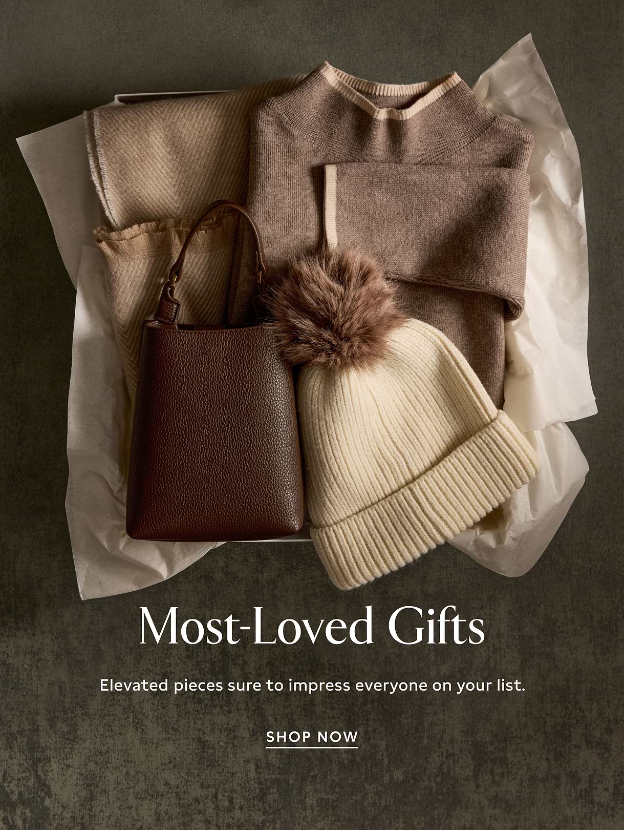 Boxed gift set with tan sweater and scarf, cream ribbed beanie with faux‑fur pom, and brown leather handbag; text reads “Most‑Loved Gifts.”