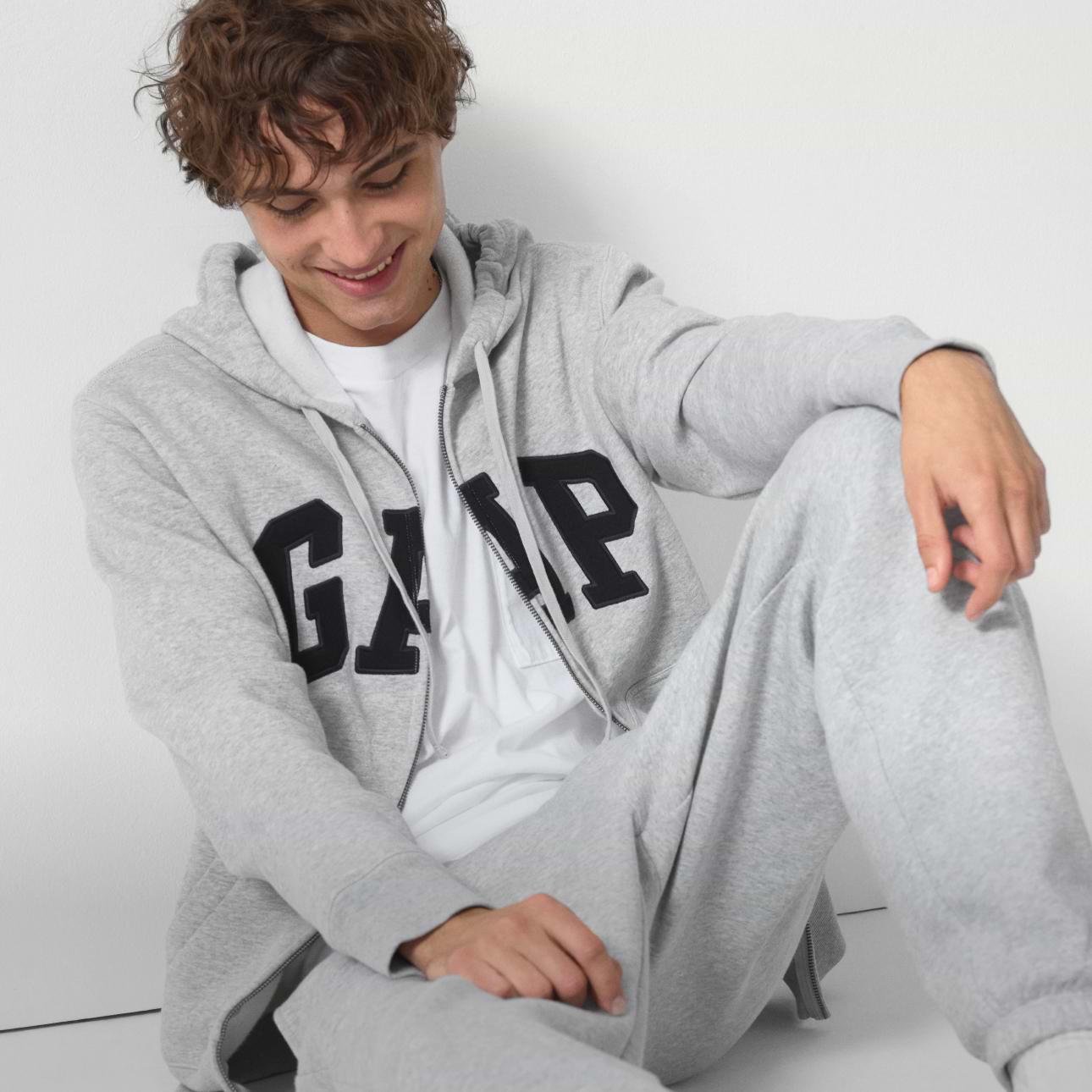 Smiling person wearing a light gray GAP hoodie and matching sweatpants, seated against a white wall.