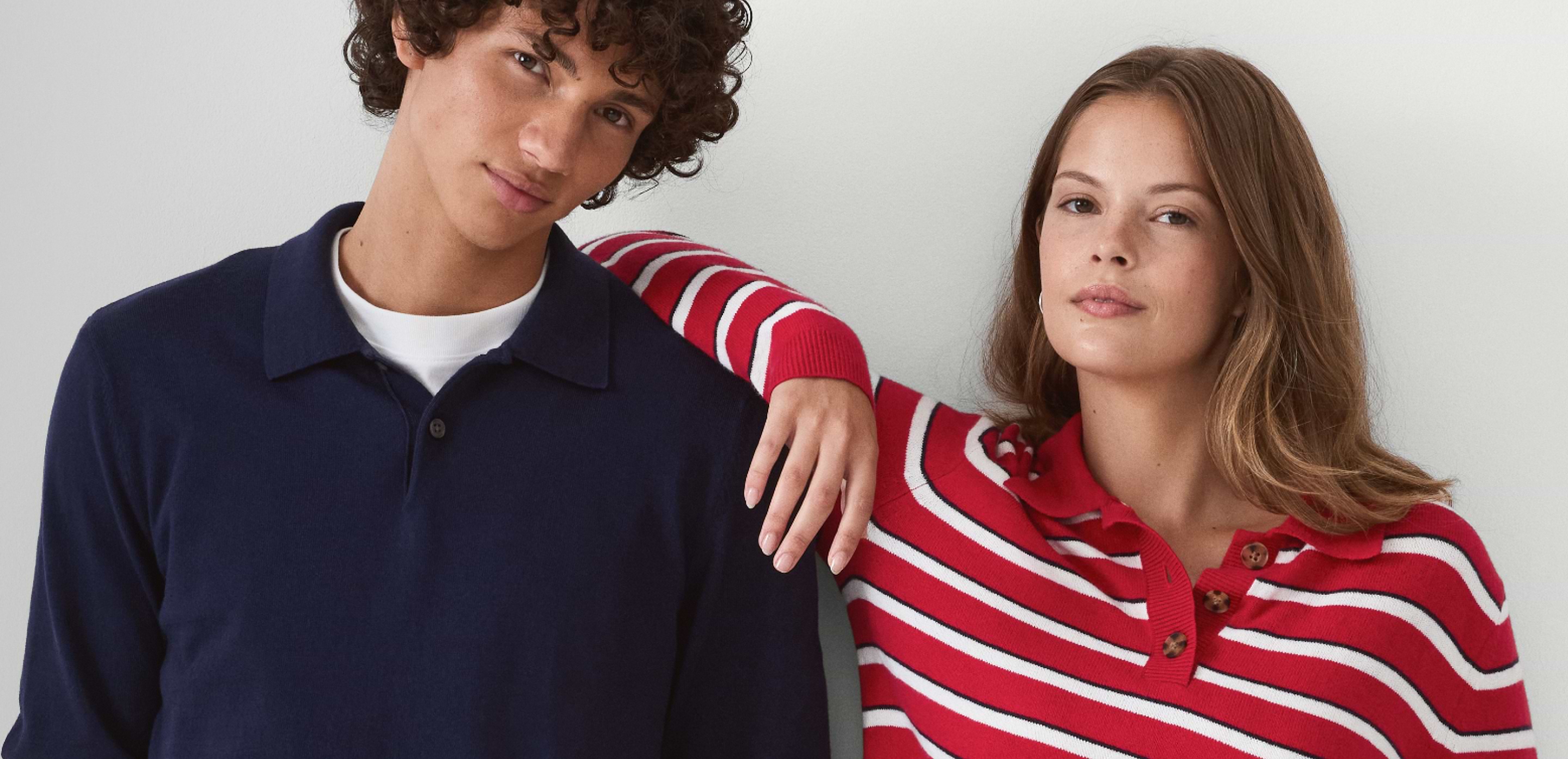 Two people pose against a light wall; the person on the right rests their arm on the other’s shoulder. Left wears a navy collared sweater, right wears a red-and-white striped collared sweater.