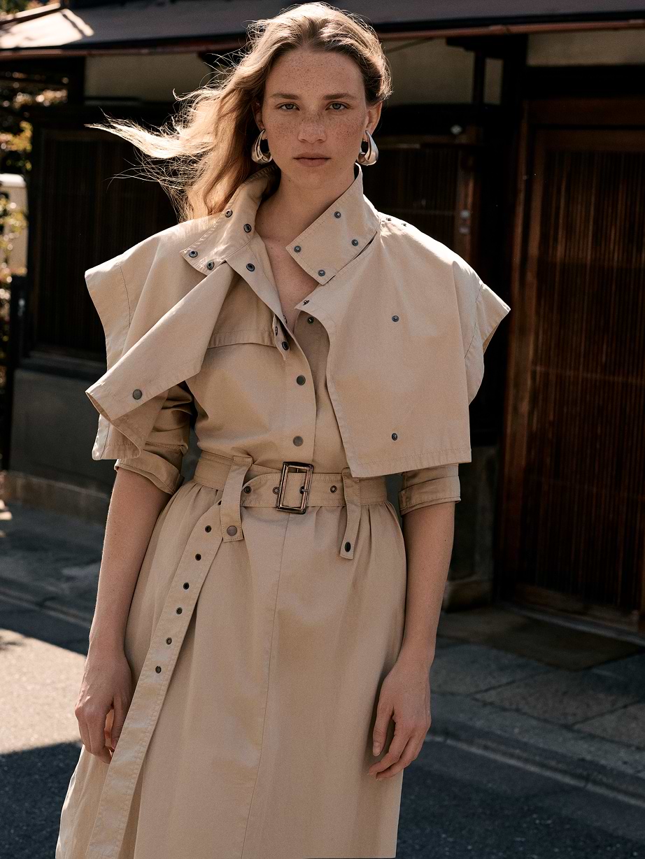 Person in a beige belted trench-style dress with cape detail, standing on a sunny street, hair blowing.