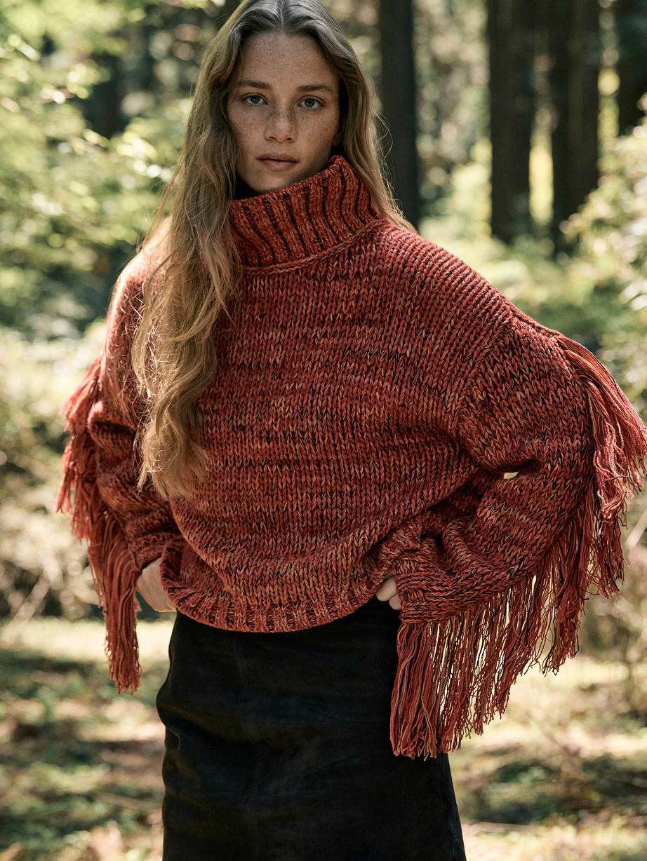 Person in a rust-colored chunky turtleneck sweater with fringe sleeves, standing in a sunlit forest.
