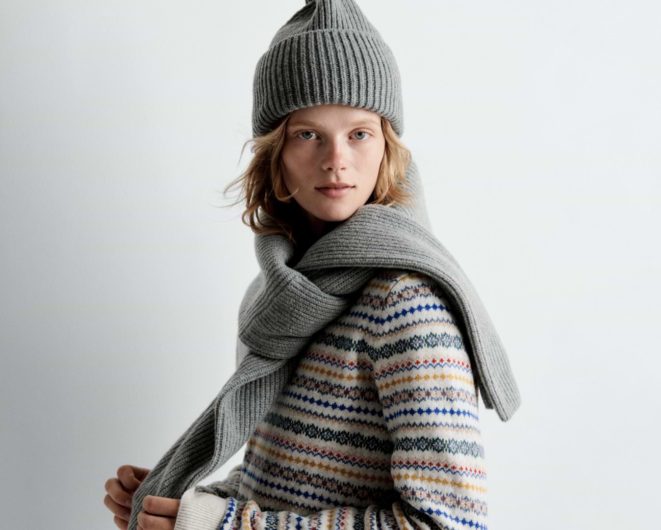 Person in a gray knit beanie and matching scarf, wearing a multicolored patterned sweater, looking at the camera against a plain background.