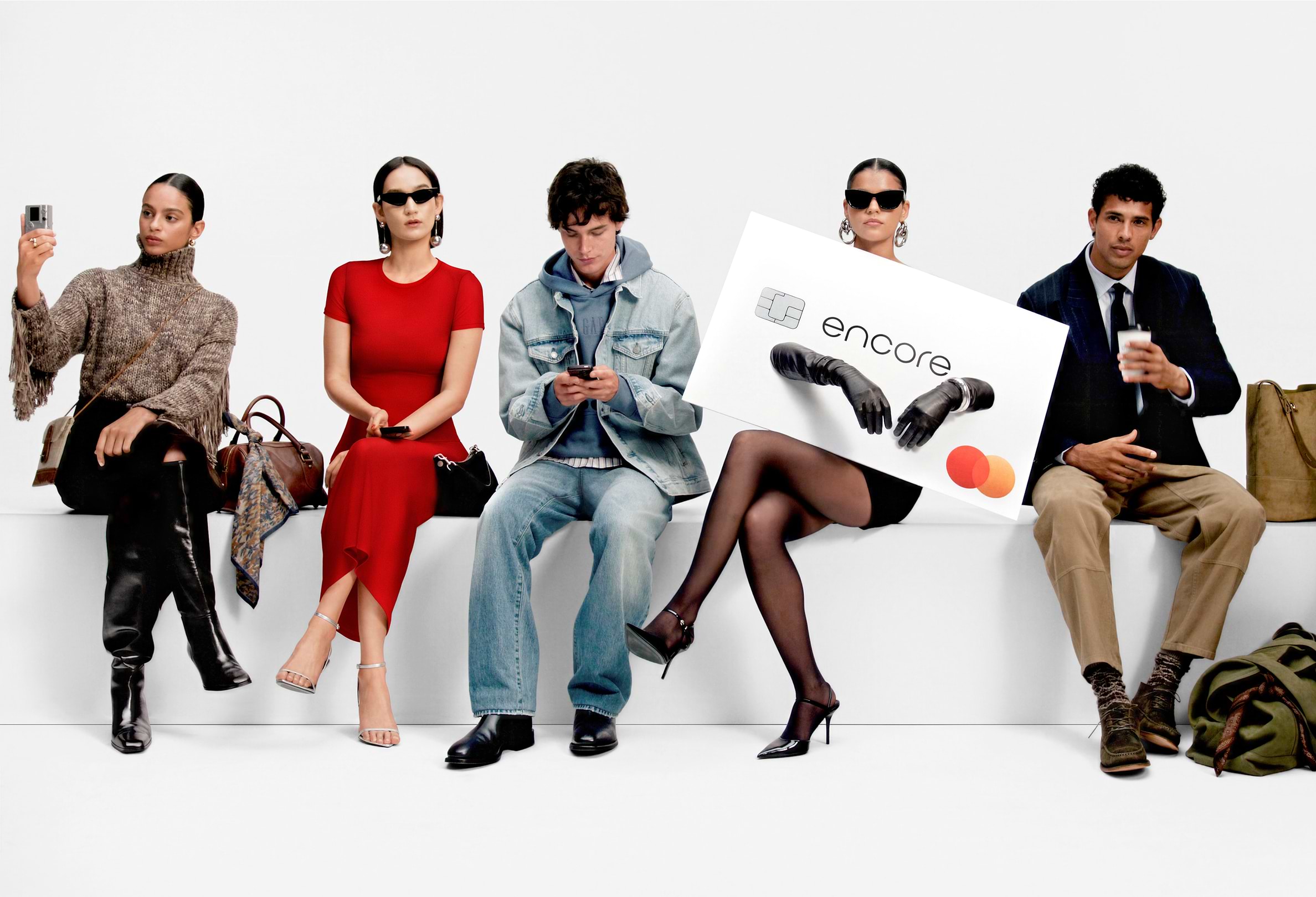 Five stylish people sit on a white bench; two use phones, one in a red dress, one in denim; another holds a large “encore” card with Mastercard logo; a man holds coffee.