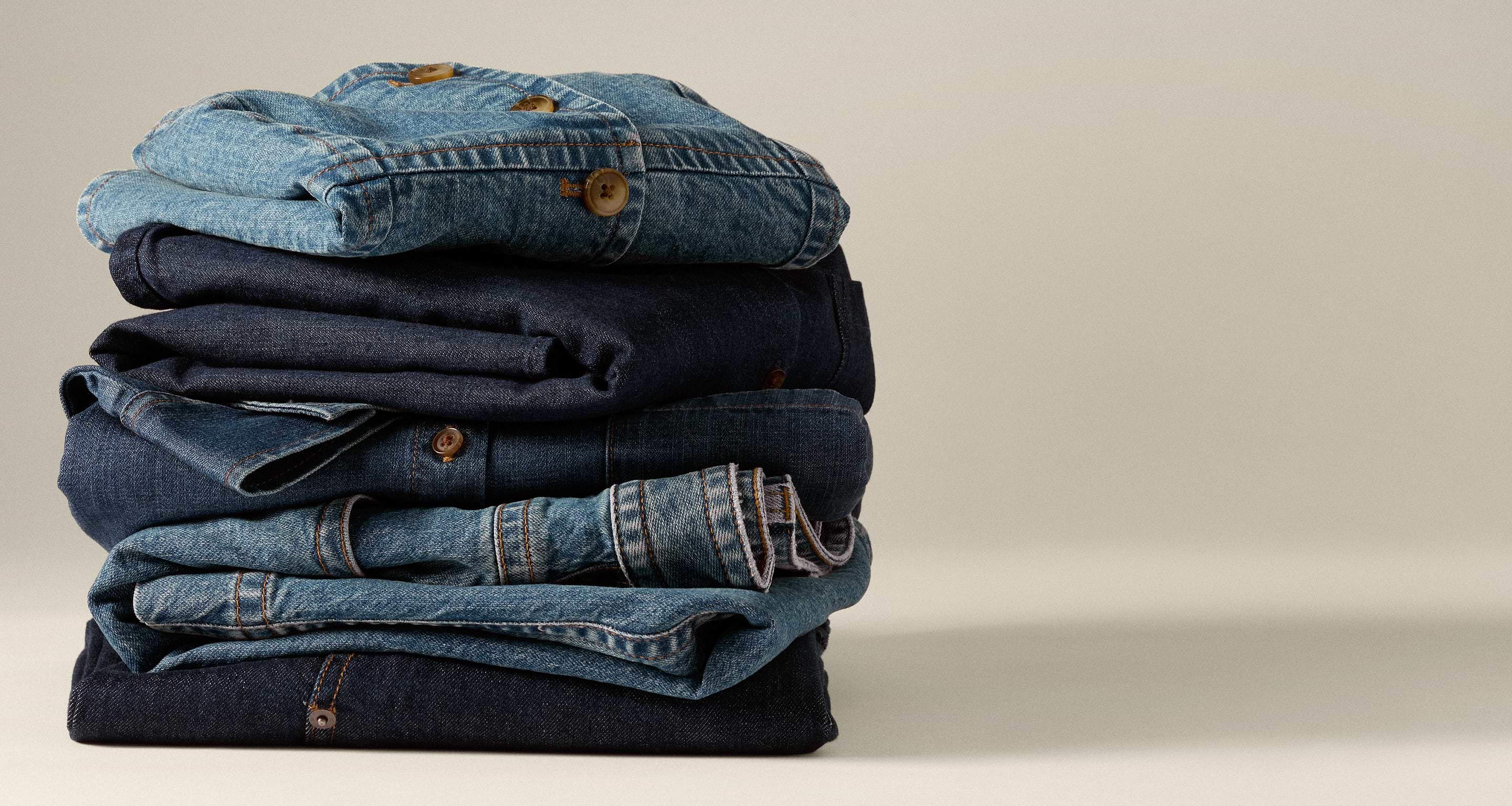 Stack of folded blue denim jeans topped with a denim shirt on a beige background.