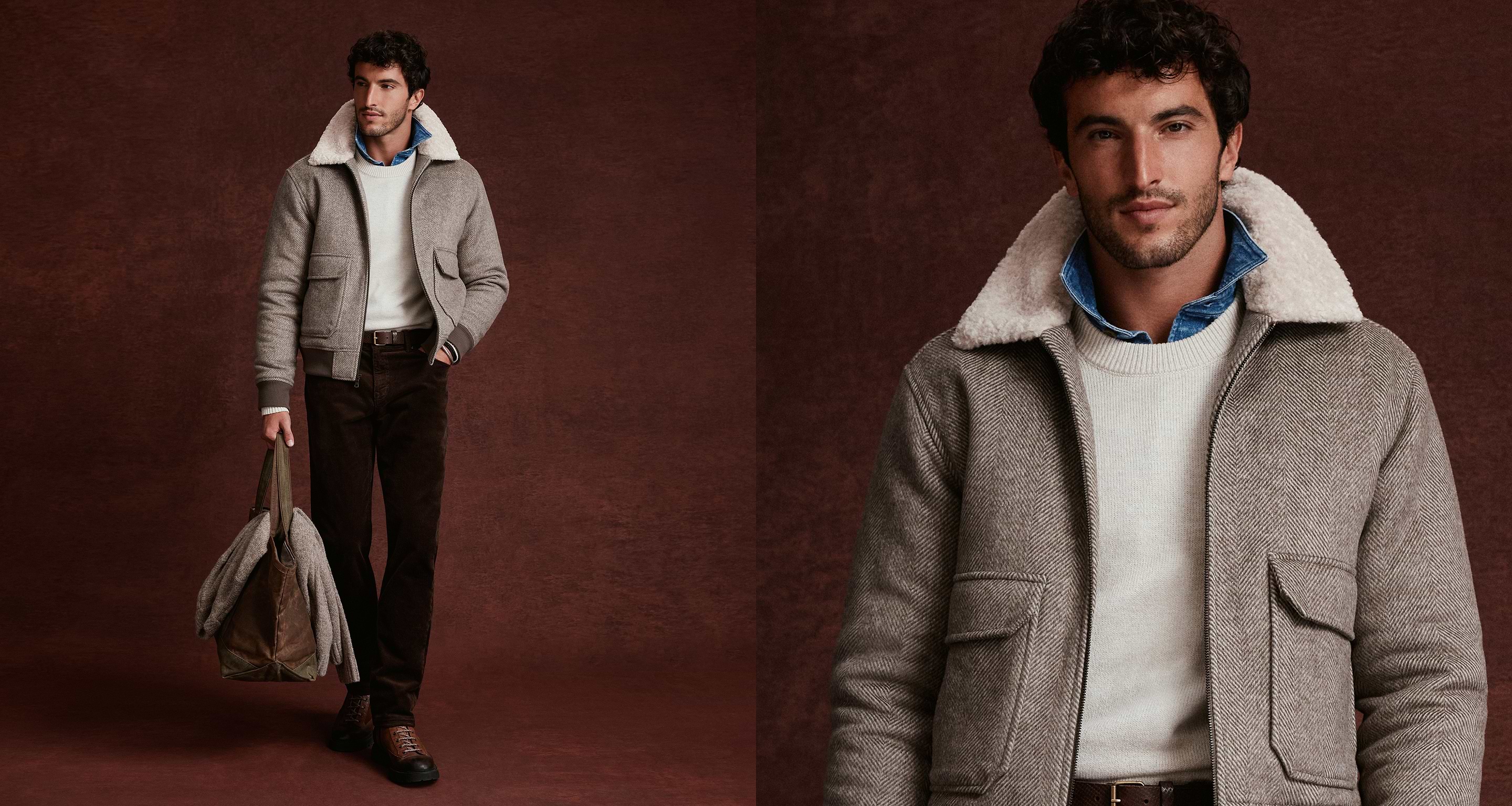 Man wearing a gray shearling-collar jacket over a cream sweater and denim shirt, dark pants and boots, holding a brown bag.