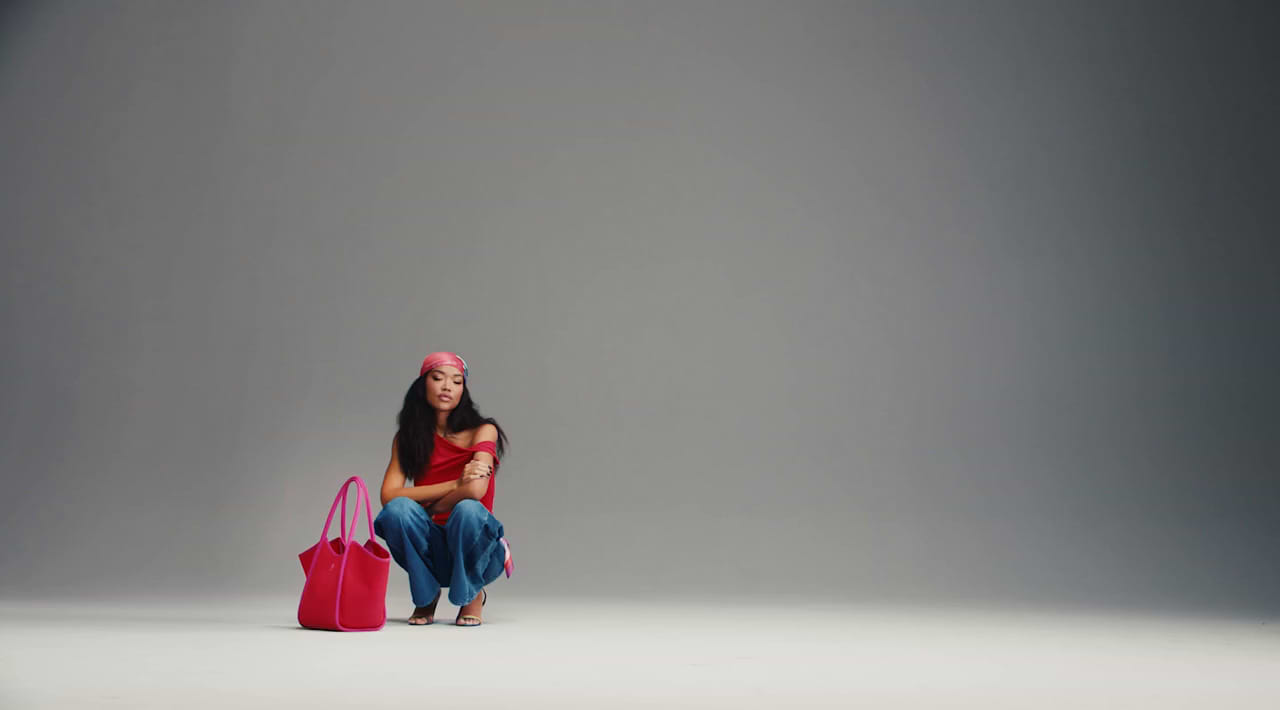 Person in a red top and headscarf crouches beside a large pink tote bag against a gray studio backdrop.