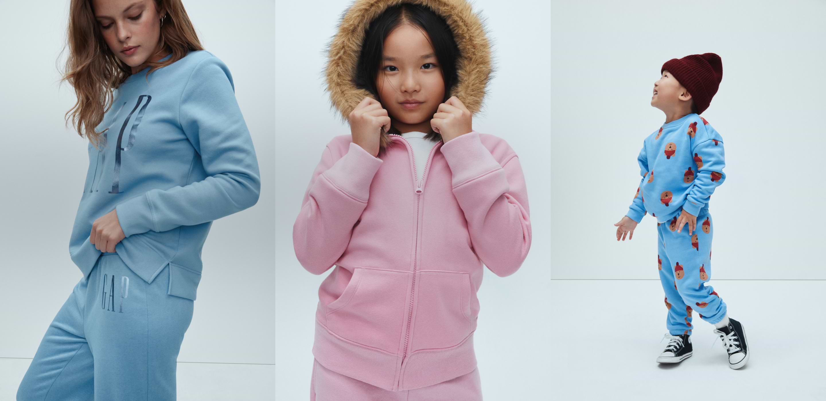 Three models in cozy loungewear: light‑blue sweatshirt set, a pink zip hoodie with faux‑fur hood, and a toddler in blue printed sweats with a maroon beanie.