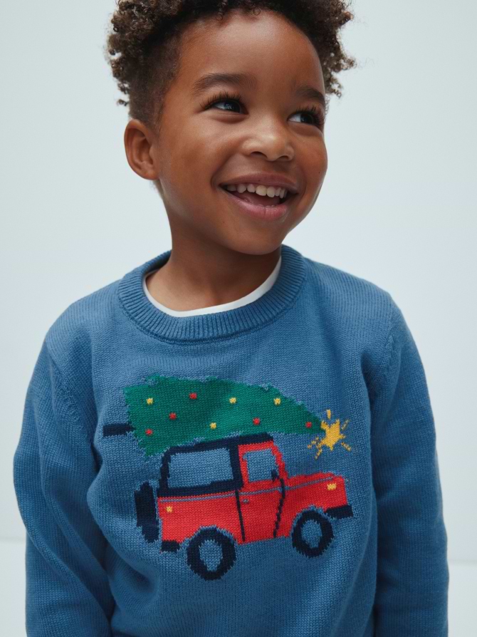 Smiling child wearing a blue sweater with a red car carrying a decorated Christmas tree design.