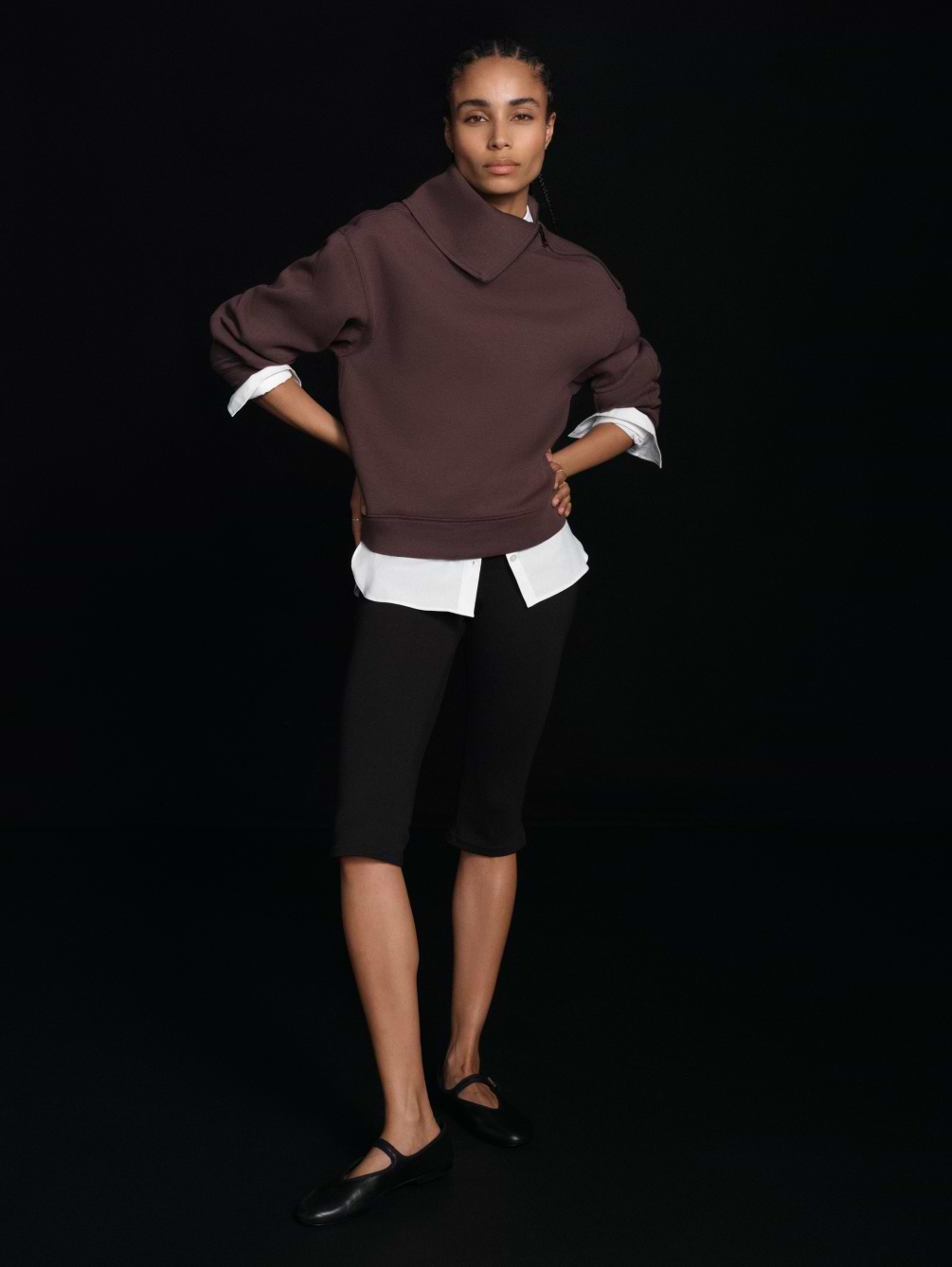 Person stands against a black background, hands on hips, wearing a brown wide‑collar sweater over a white shirt, black knee-length leggings, and black flats.