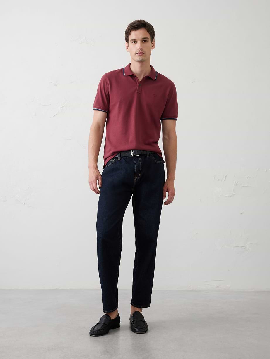 Person standing in a studio wearing a maroon polo shirt, dark blue jeans with a belt, and black loafers against a plain white wall.