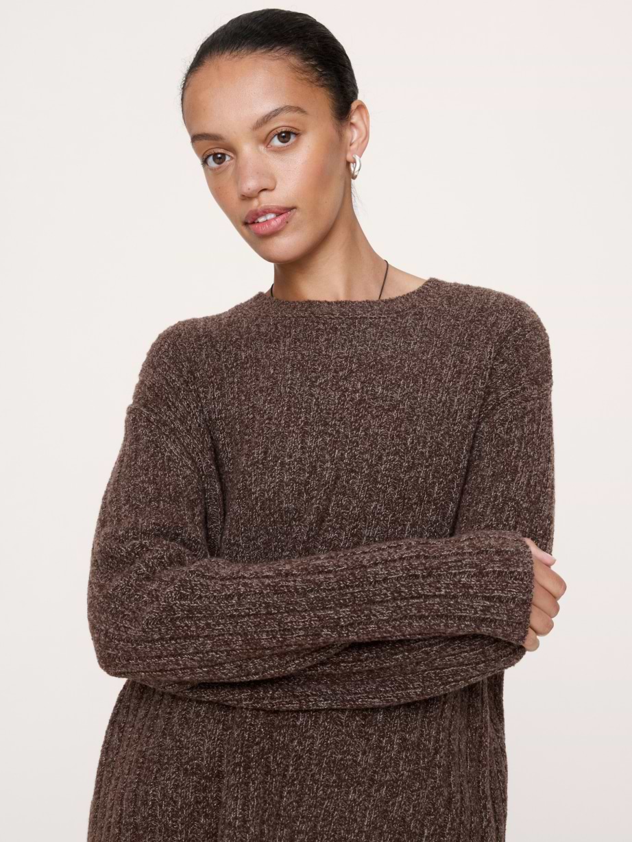 Woman in a brown ribbed knit crew‑neck sweater with arms crossed against a light background.