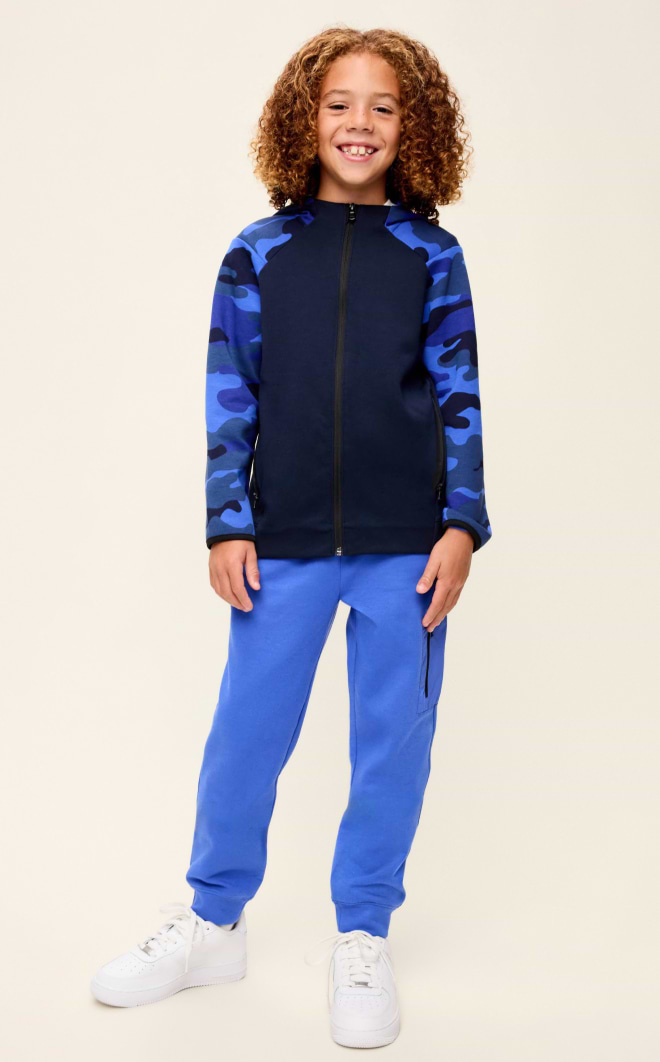 Smiling child in a navy zip-up jacket with blue camo sleeves, bright blue joggers, and white sneakers, standing against a plain background.