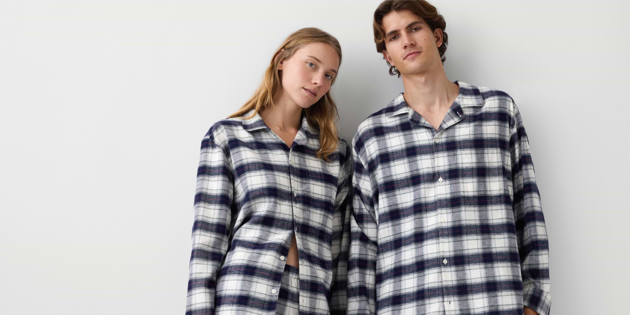 Two people in matching white-and-navy plaid flannel pajama shirts stand side by side against a plain wall.
