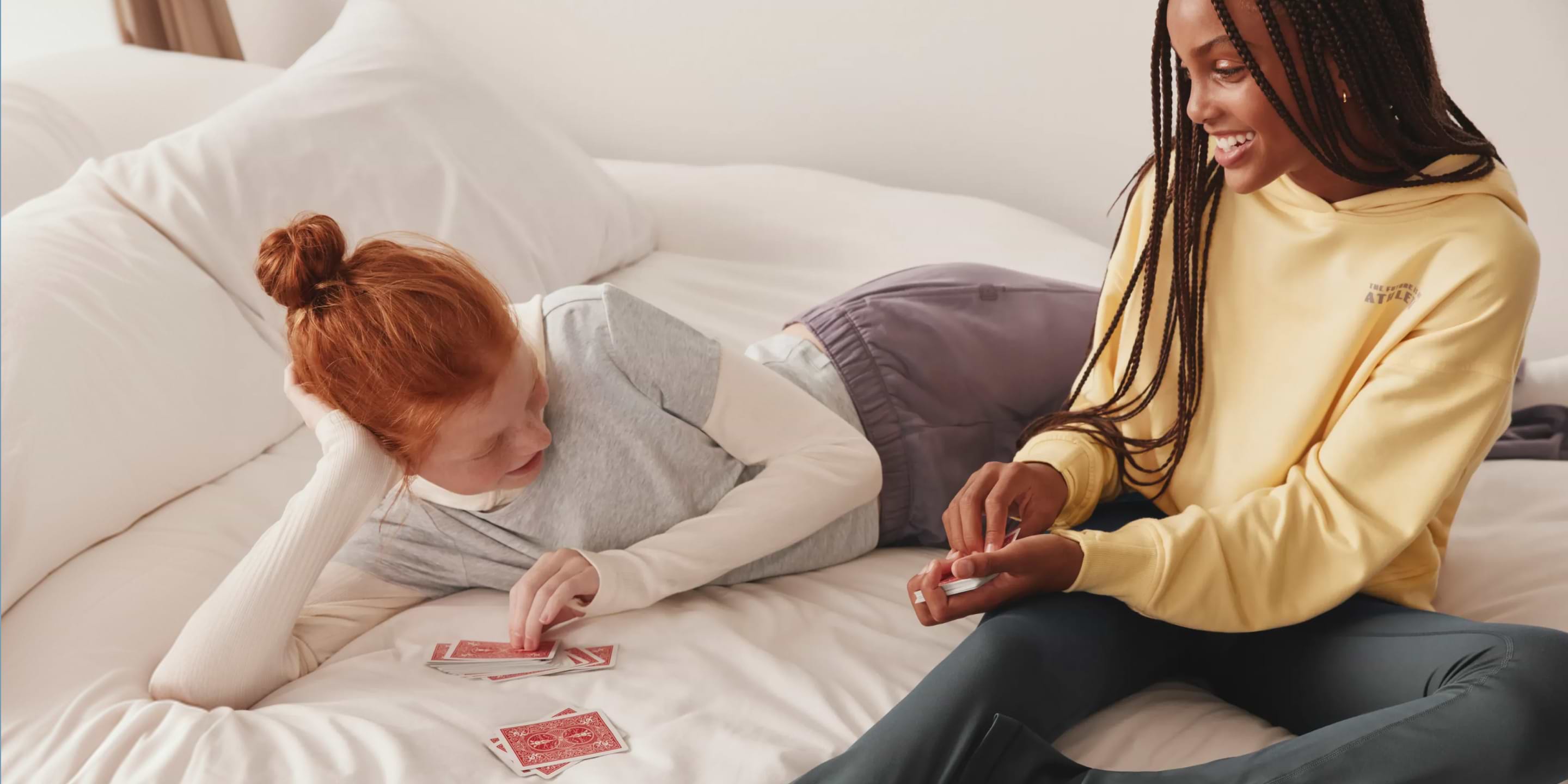 Two girls relax on a bed, smiling and playing cards—one lying on their side while the other deals.