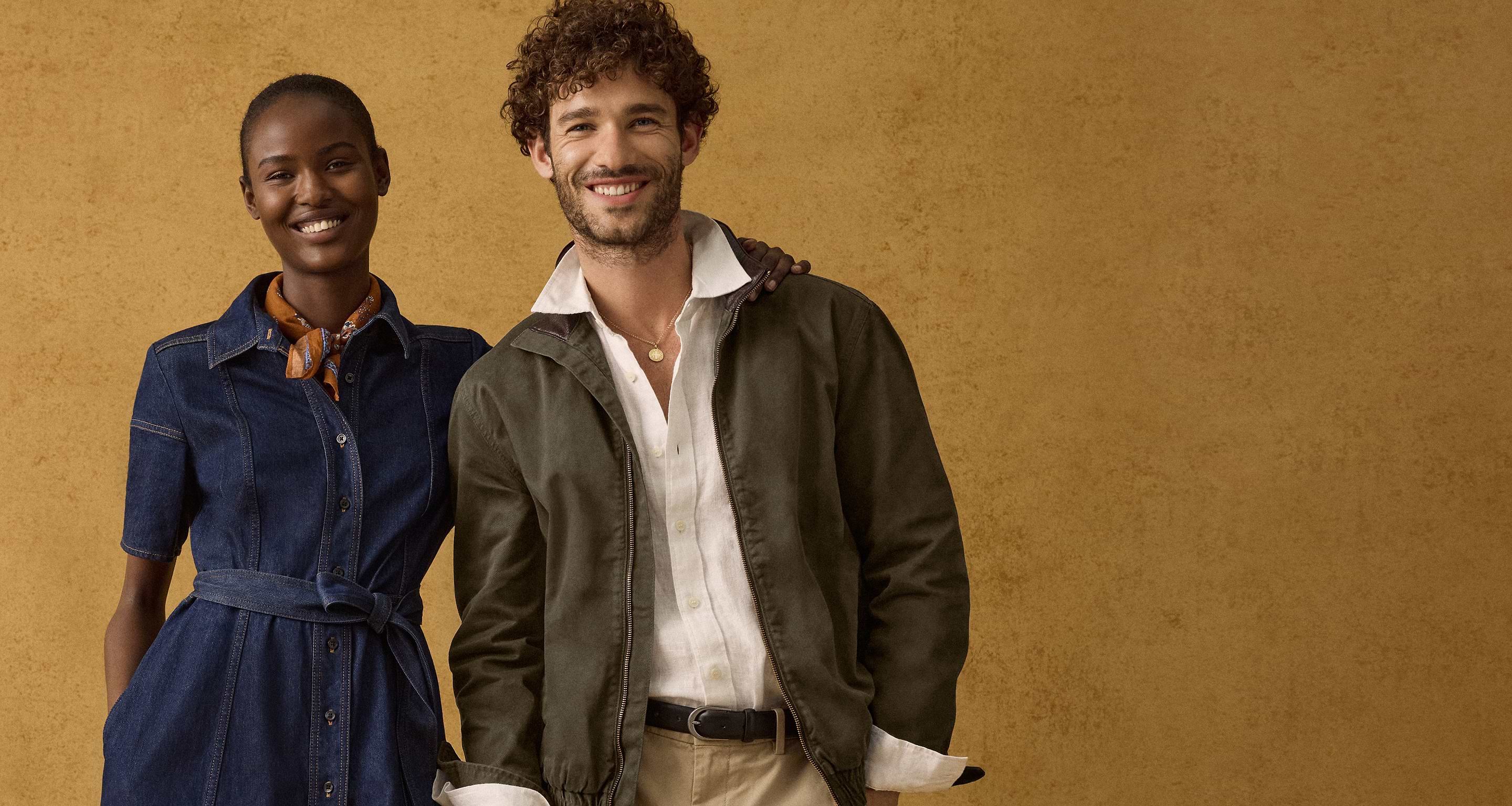 Two smiling models against a tan backdrop: woman in a belted denim dress with scarf, man in an olive jacket over a white shirt and khaki pants.