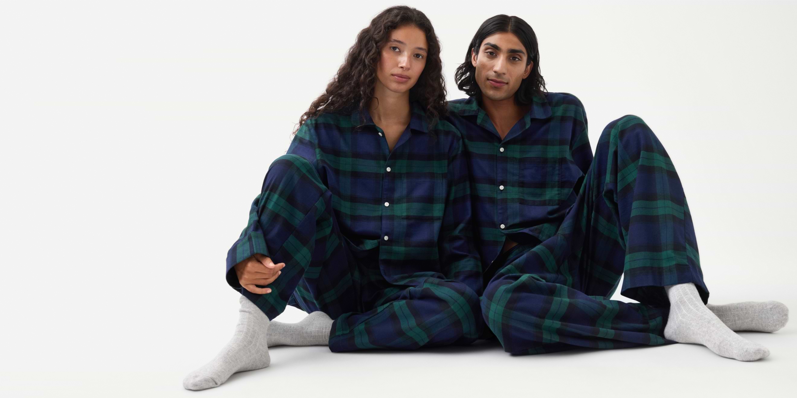 Two people sit on the floor in matching navy-and-green plaid pajamas and gray socks against a plain white background.