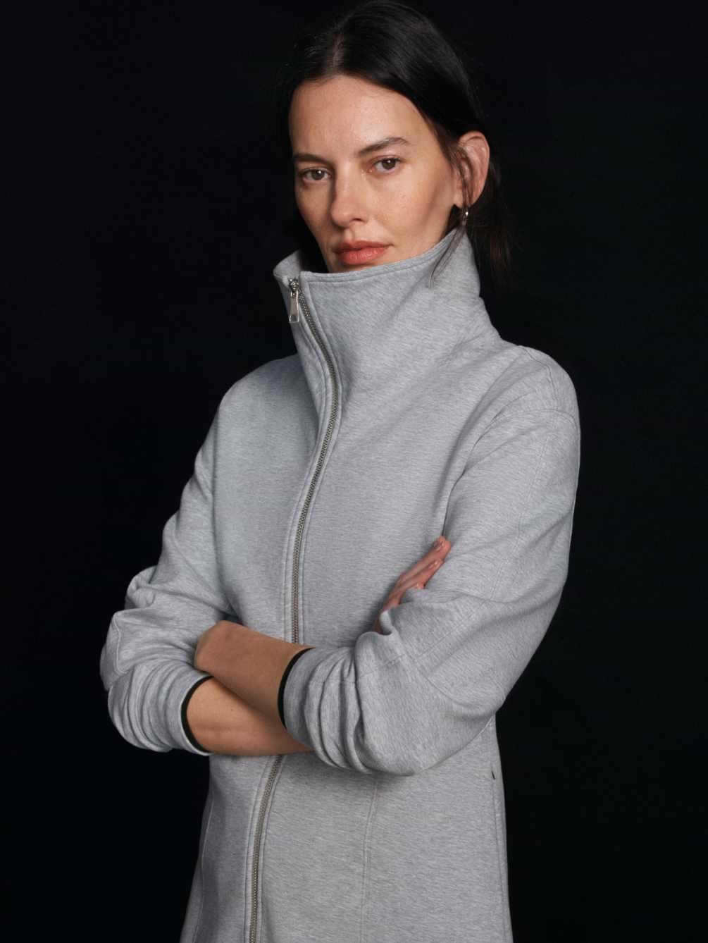 Woman in a light gray zip-up jacket with a high collar, arms crossed, against a black background.